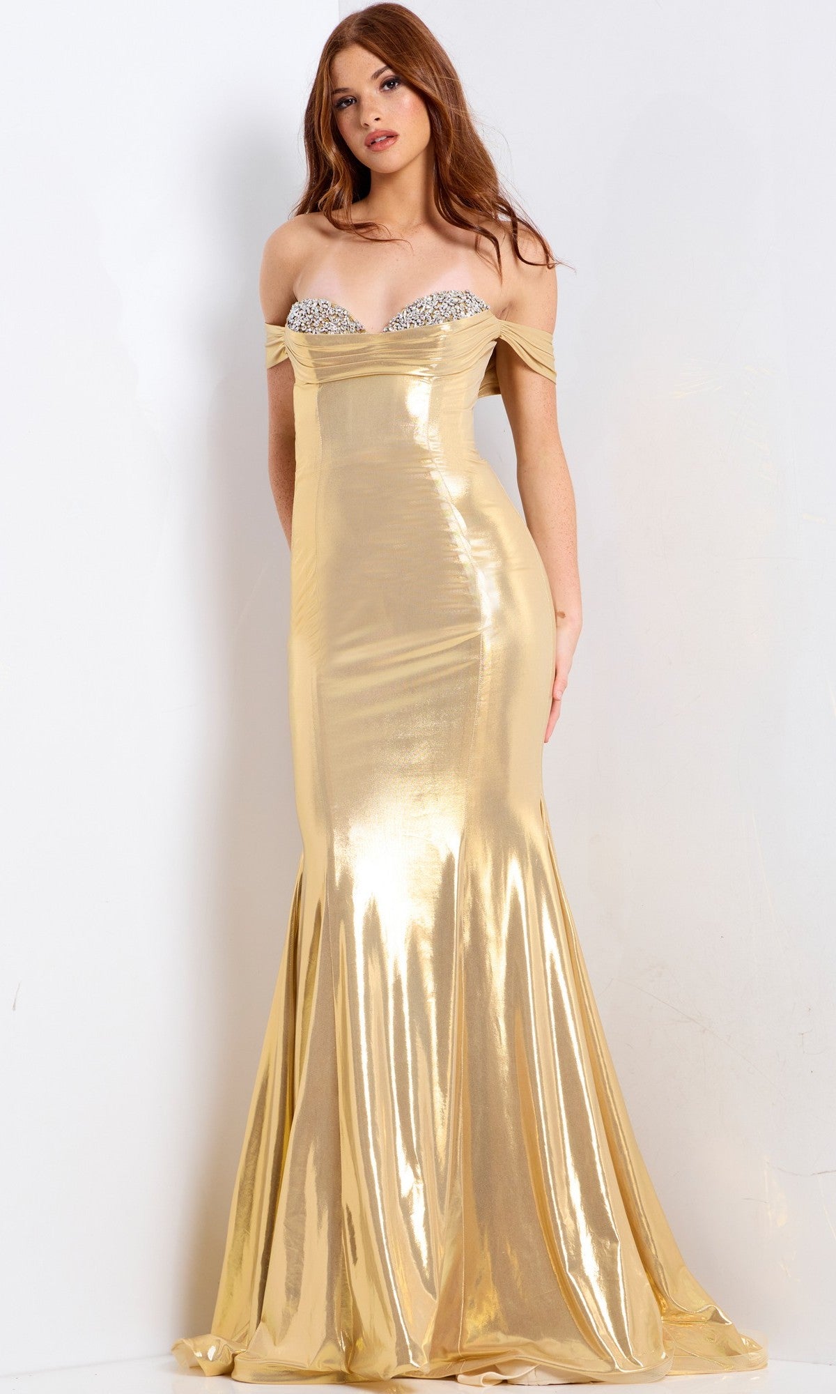 A woman stands against a plain white background wearing the Jovani 47153 long prom dress, featuring shiny gold metallic fabric, a fitted bodice, an embellished sweetheart neckline, and a floor-length skirt.